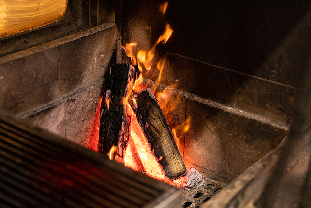 肉を焼く１時間半ほど前から火を熾し始める。最初は火床が冷たいので熾火になるまで時間がかかるが、営業が進むにつれ火床が温まり、60～80分で熾火になる。空気が通る隙間を作るように薪を立てて燃やす。