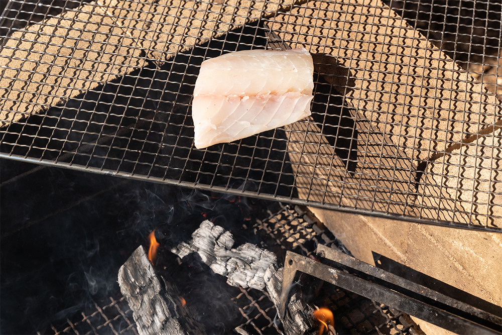 火を消して煙が立つ状態にした薪から20cm上に網を置き、クロダイの切り身をのせる。