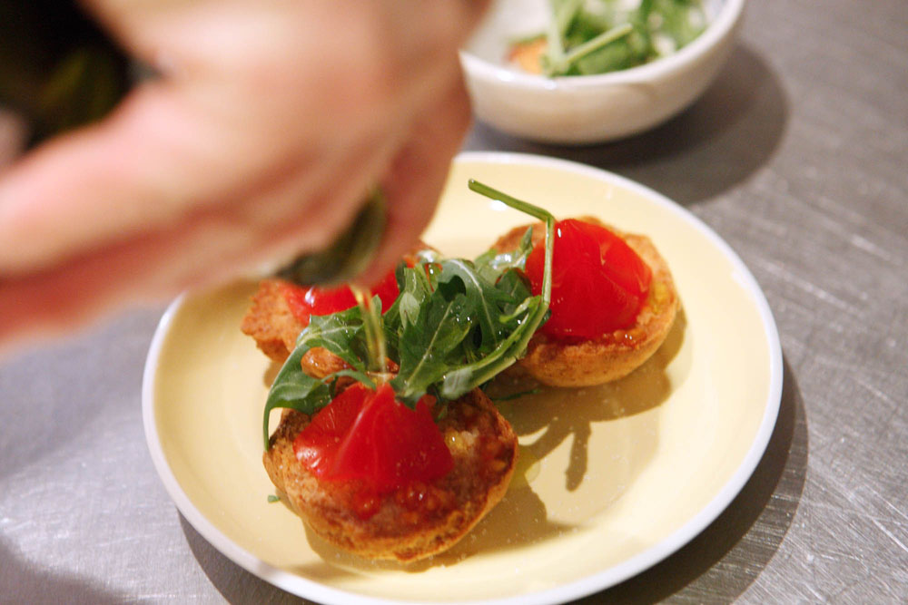 [３] トマトを表面にこすりつけてのせる。ルッコラ（バジルでも可）を飾り、塩をふり、オリーブオイルを回しかける。